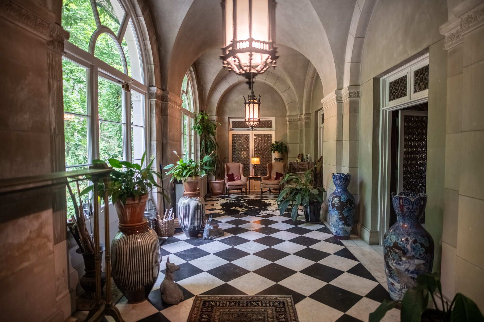 A vaulted stone gallery with arched windows, a checkerboard marble floor, tall blue-and-white vases and potted palms.