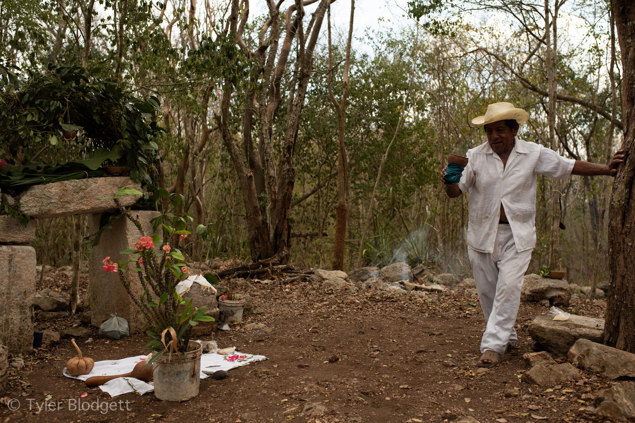 An elder in white and a straw hat walks up to the forest altar, a censer in hand.