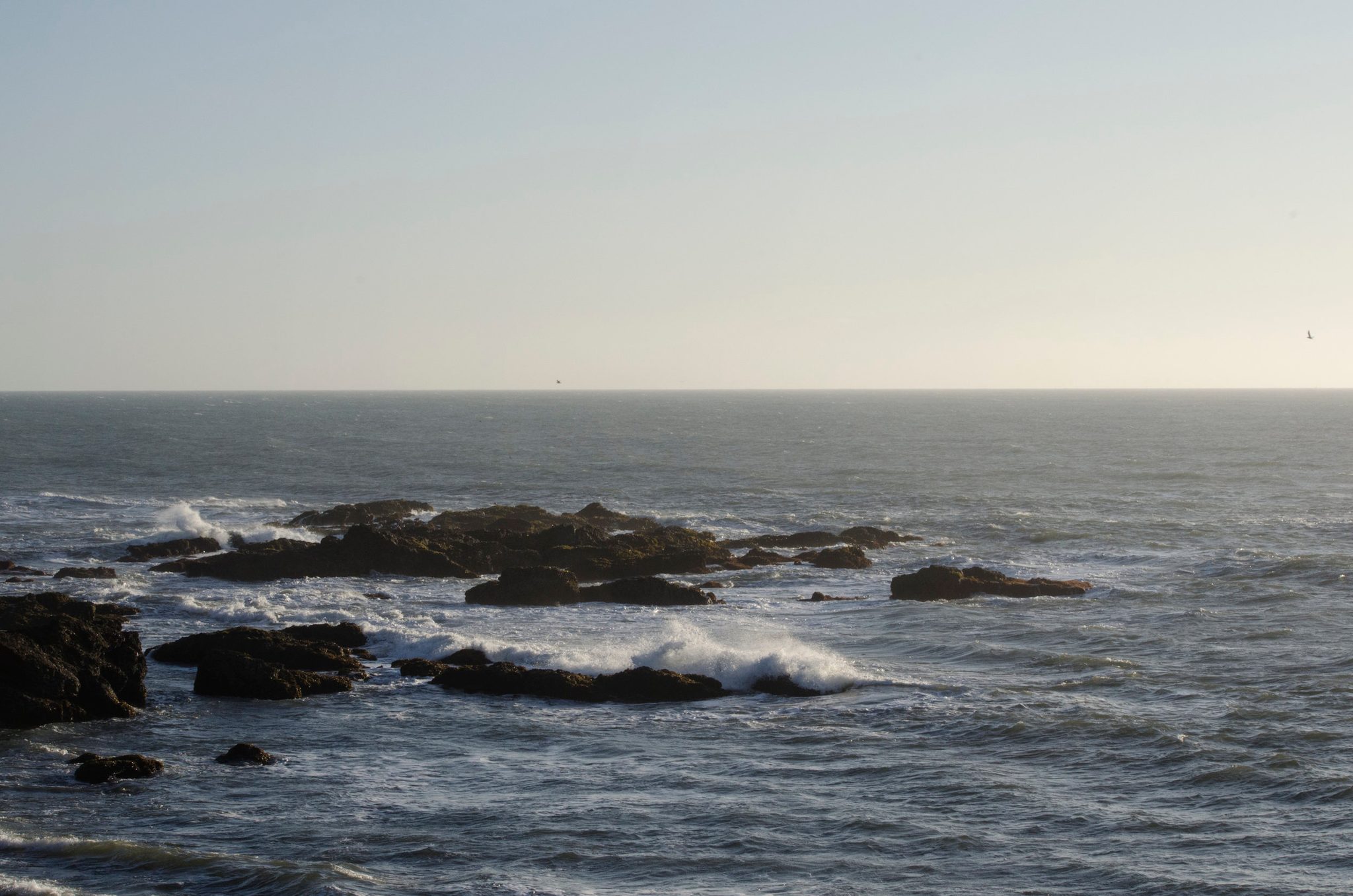 Waves break over the dark rocks under a pale sky.