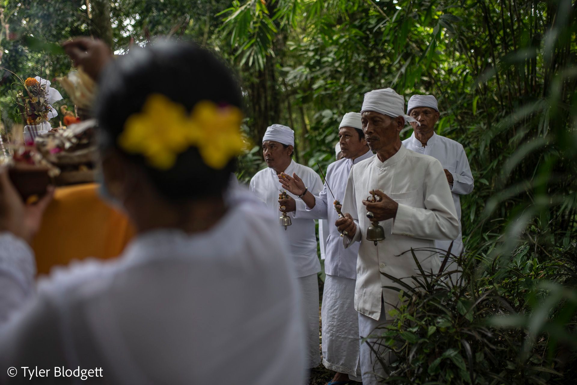 Priests ring their bells at the edge of the forest. A woman with yellow flowers in her hair carries an offering past.